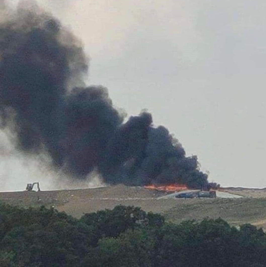 A large plume of black smoke rises dramatically from a hillside, indicating a possible fire or explosion in the area